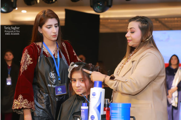 A salon or hair styling event with three women in the foreground. One woman on the left wears a red embroidered robe over a black apron, while the woman on the right in a beige blazer styles the hair of the seated woman in the center.